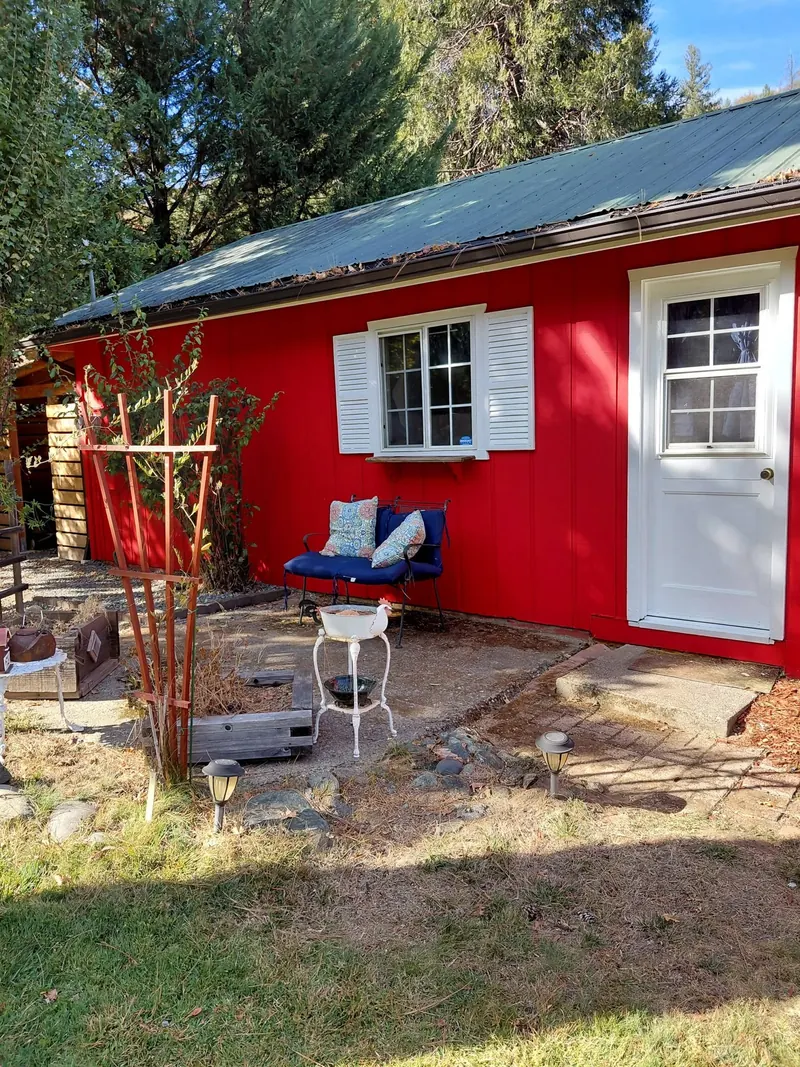 Entry side of the red home with a white door and shuttered window, a garden trellis, and a bench with blue cushions.