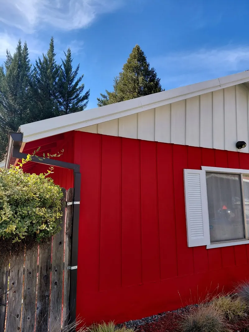 Corner of a red board-and-batten home with a white roofline, a dark gutter and downspout, and tall evergreens against a blue sky.