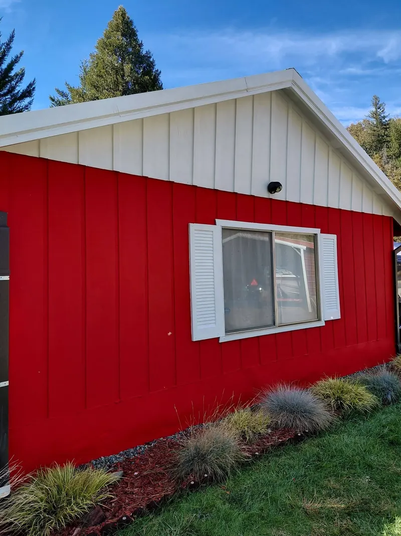 A red board-and-batten wall under a white gable, with a white-shuttered window and ornamental grasses along the base.